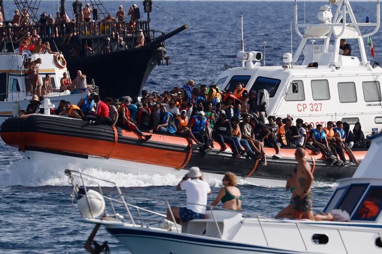 An Italian Coast Guard boat carries migrants as tourists on a nearby boat watch near the port of the Sicilian island of Lampedusa, southern Italy, Sept. 18, 2023. 