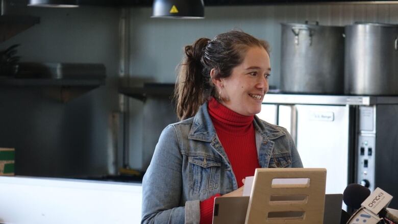 woman with hair in a ponytail wearing an orange turtleneck and jean jacket speaking to journalists