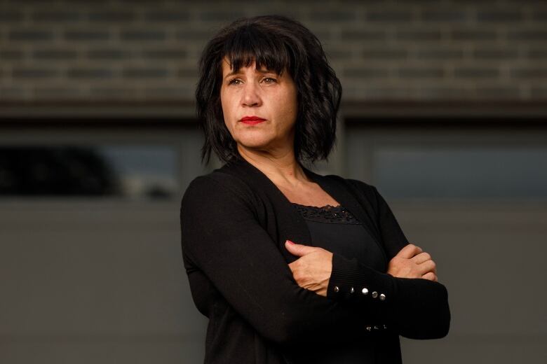 A medium close up shot of a white woman wearing a black shirt and cardigan standing with her arms crossed. 