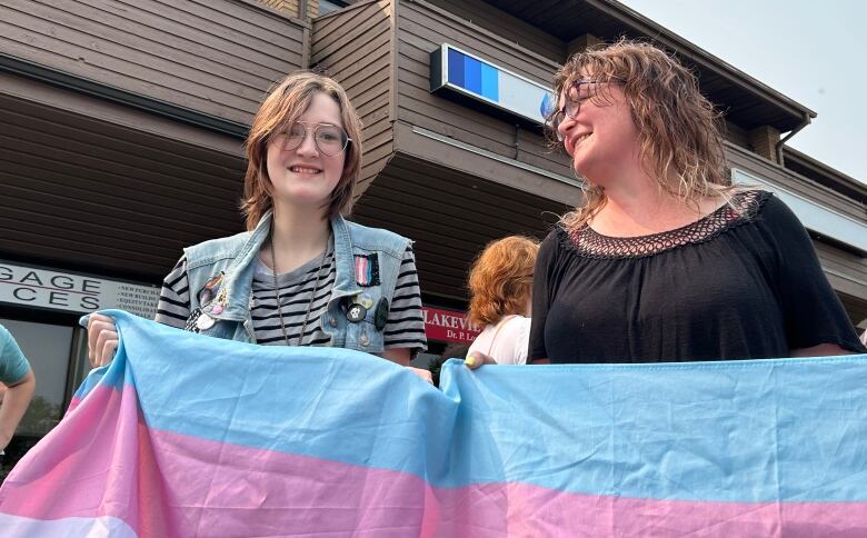 A smiling teenager and a woman stand in a crowd of people outside, holding a multicoloured transgender Pride flag.