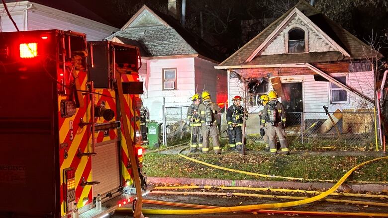 Firefighters stand on the sidewalk in front of a home. A fire truck is parked on the street.