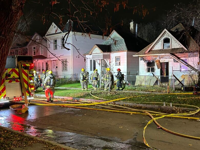 Firefighters stand on the sidewalk in front of a home. A fire truck is parked on the street.