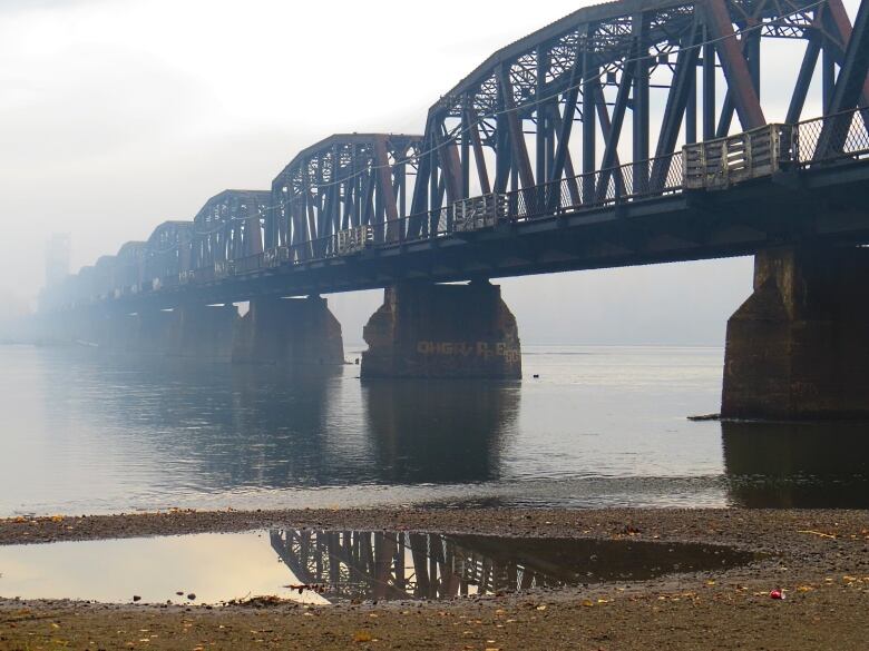 A bridge over water is seen from sandy a shore, with a small puddle of water on the shore. The bridge's far end is covered in mist.