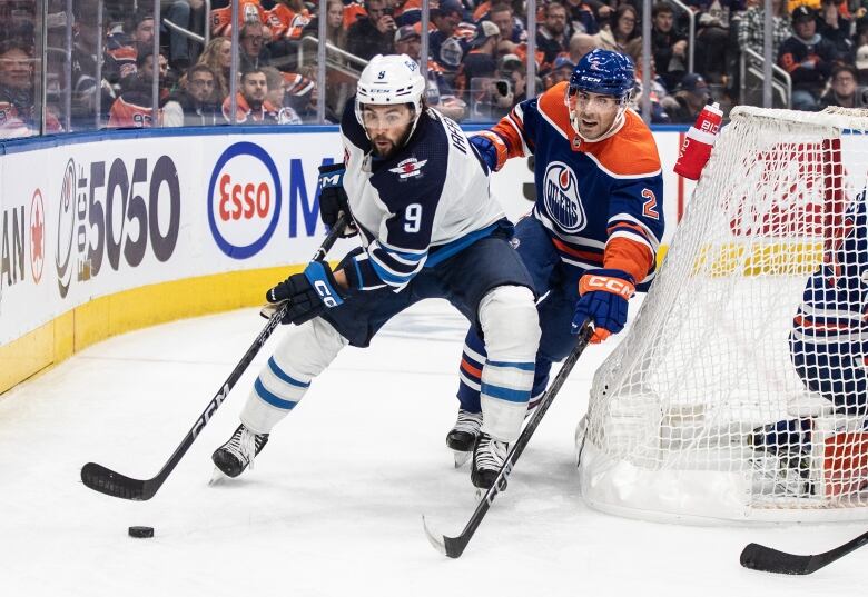 A player in a white jersey skates toward a hockey puck while a player in a blue jersey reaches his hockey stick next to him.