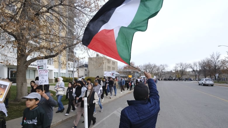 A man in the foreground with his back to the camera waves a Palestinian flag. In the background, people march down the sidewalk.