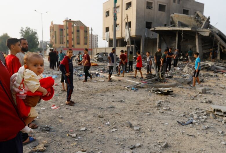 A person holds a baby as people gather at buildings in ruin.