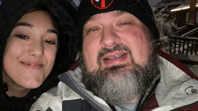 A teenage girl and her father smile for a selfie. They are dressed warmly, standing outside in winter with snow-covered trees in the background.