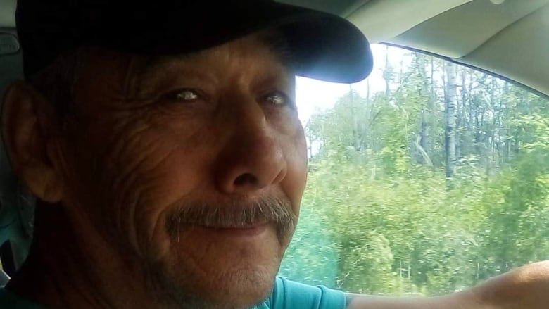 A man wearing a ball cap is seen sitting in a vehicle, with trees in the background, and looking at the camera.