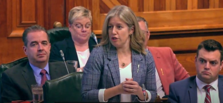 A woman wearing a blazer and blouse, gesturing with her hands as she speaks into a microphone, with four people behind her looking on.