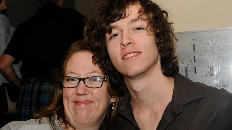 A middle aged woman leans into the crook of the arm of a teenage boy. Both are smiling at the camera.