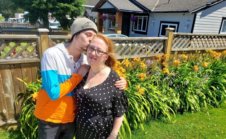 A teenage boy kisses a middle-aged woman on the cheek while they stand in a garden. The woman is smiling.