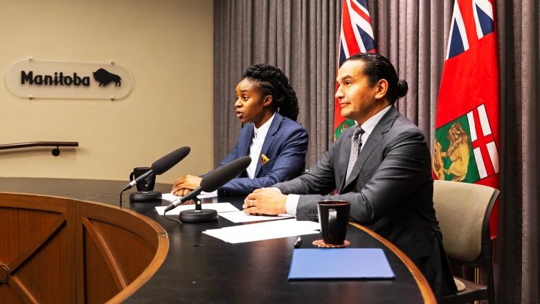 Two people in blazers are seated at a table, speaking with members of the media.