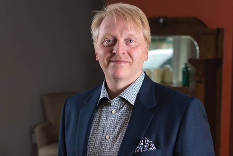 A man in his forties or fifties with strawberry blonde hair and wearing a blue blazer and white and blue dress shirt with a square and dot pattern smiles with his mouth closed for a head and shoulders portrait. Part of a mantle, mirror and antique chair can be seen in the background.