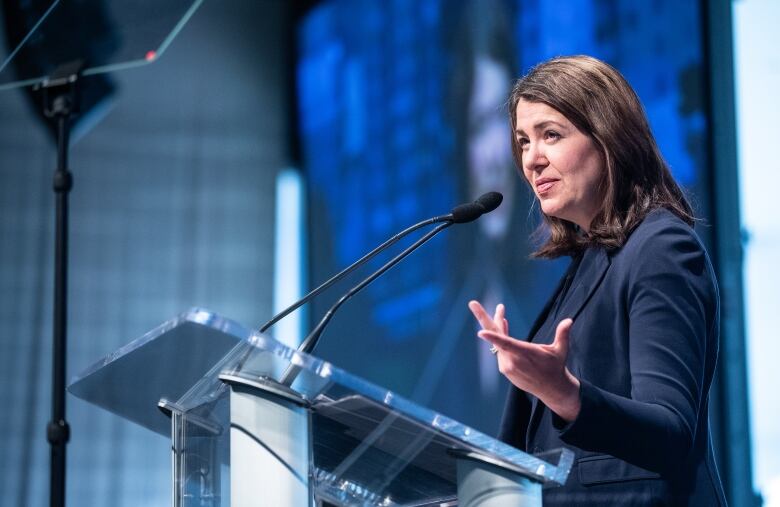 A woman wearing a dark blue blazer standing on a podium, giving a speech on a stage with a blue background. 