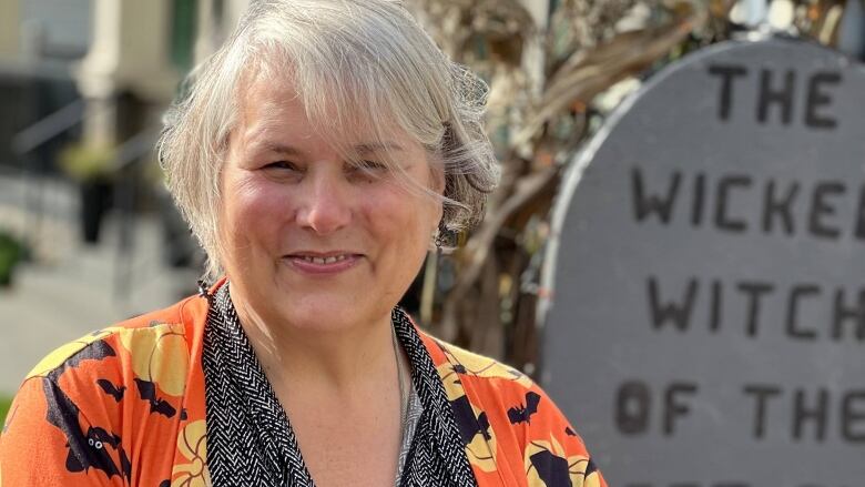 Suzanne Parnell smiles at the camera standing on her front lawn. Behind her a tombstone reads The Wicked Witch of the West End.