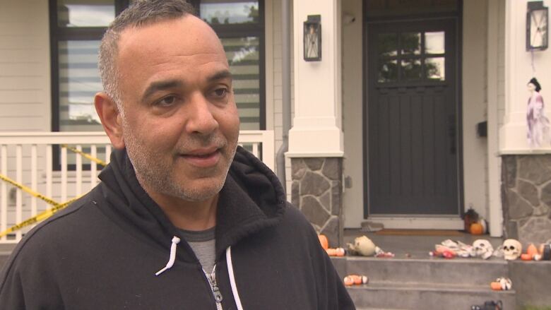 Kam Shergill stands on his front lawn smiling. Behind him decorative skulls rest on the front porch.