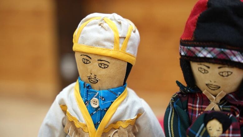 An Innu tea doll has a white hat and jacket and blue plaid shirt. 