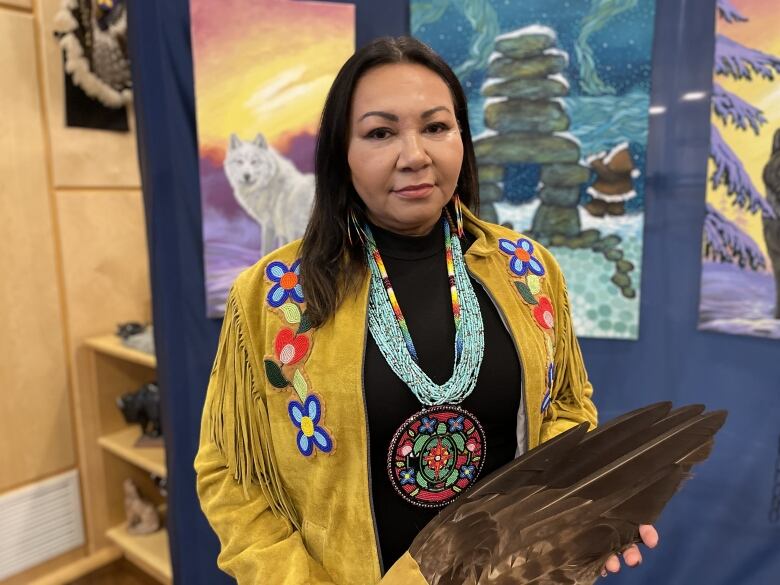 An Indigenous woman with mid-length, dark brown hair, is pictured smiling to the camera. She is wearing a beaded medallion and leather fringe jacket.