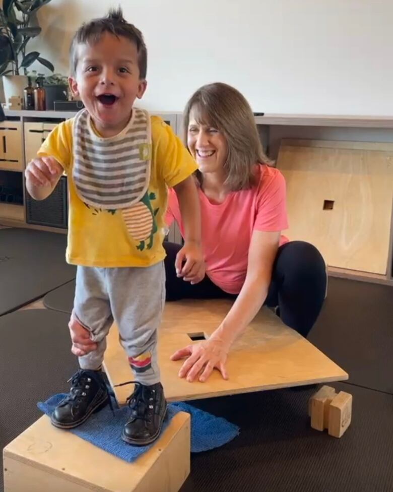 A boy stands on a wooden block with his physiotherapist behind him.