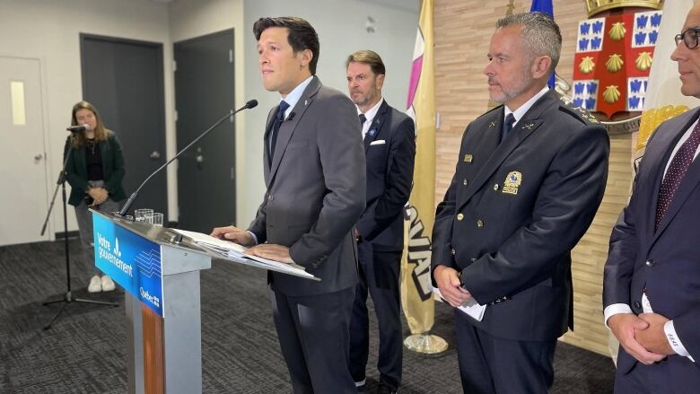 A man at a podium. Behind him there is another man in suit and a police officer in dress uniform.