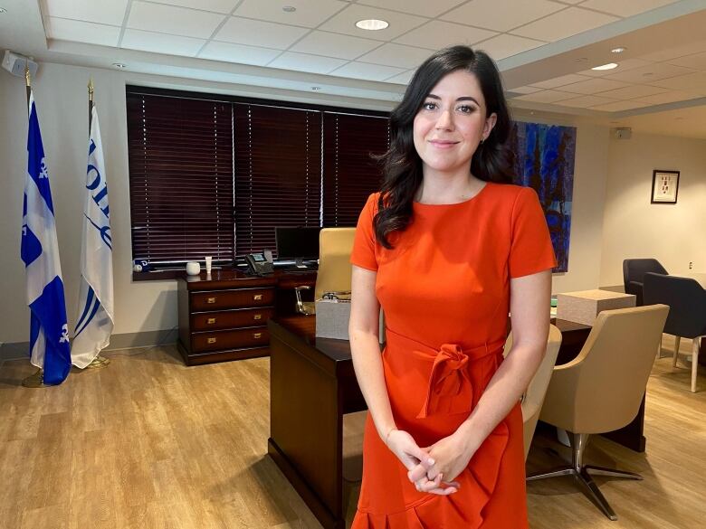 A woman in a red dress stands in front of a large desk and a few chairs.