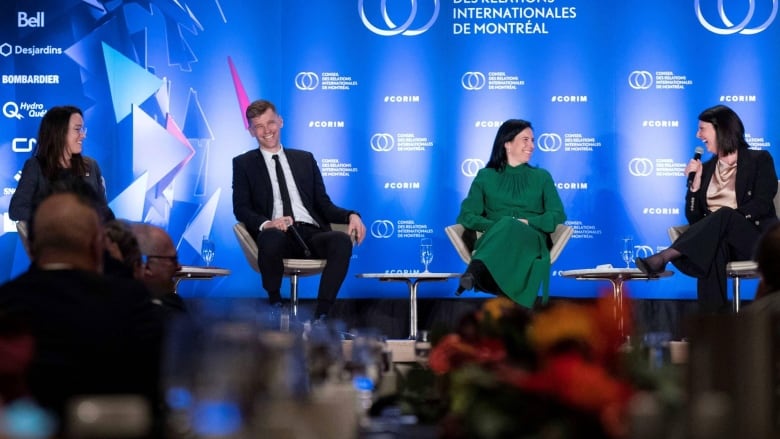 Three women and a man in business attire sit in chairs on a stage with a blue backdrop.