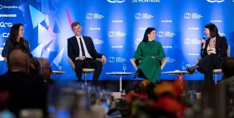 Three women and a man in business attire sit in chairs on a stage with a blue backdrop.