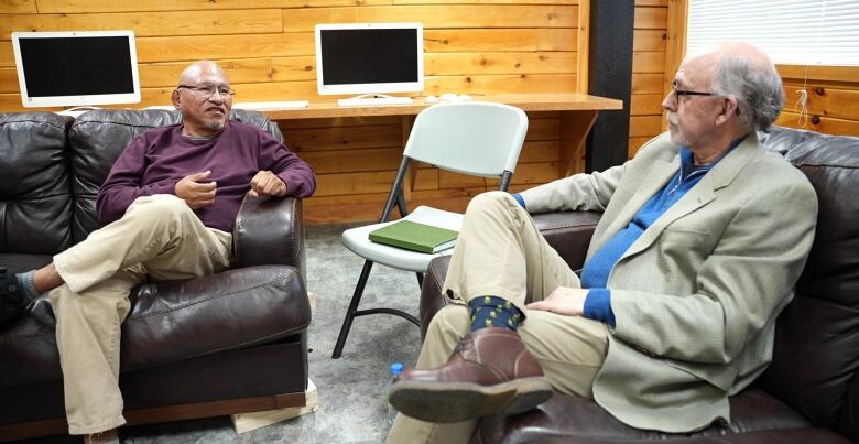 Two men sit on a black couch. One is wearing a purple sweater while the other is wearing a beige suit. 