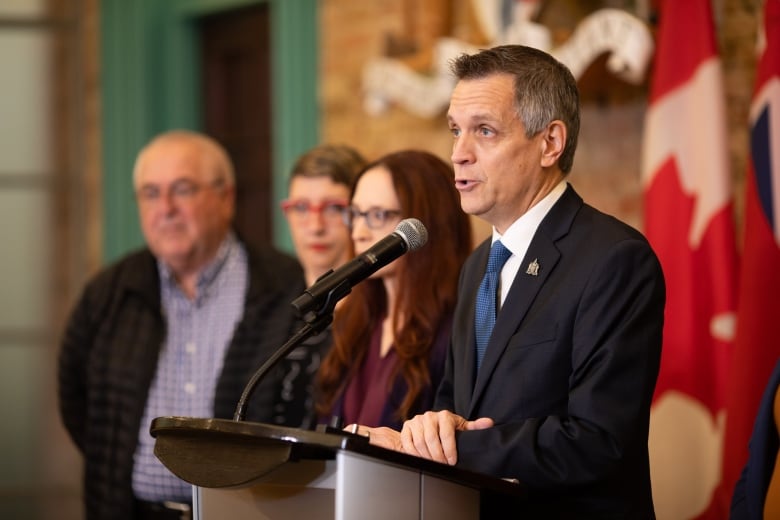 Four politicians at an announcement. One of them is speaking at a microphone.