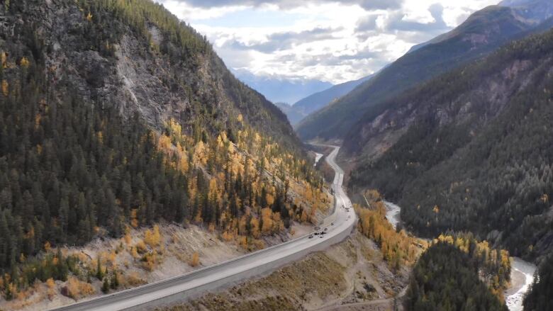 A stretch of paved highway is seen zig-zagging between mountains against a bright, cloud-filled blue sky. 