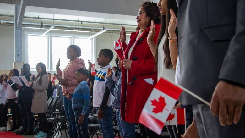 A group of people stand holding Canadian flags raising their hands for a citizenship oath.