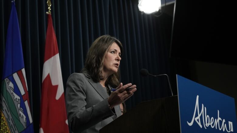 A woman gestures from behind a podium with the flags of Alberta and Canada on the stage behind her.