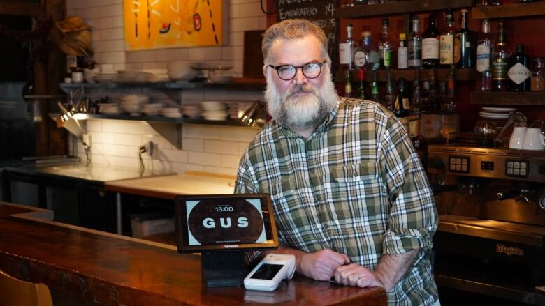 Restaurant owner with plaid shirt and white beard leans against his bar. 