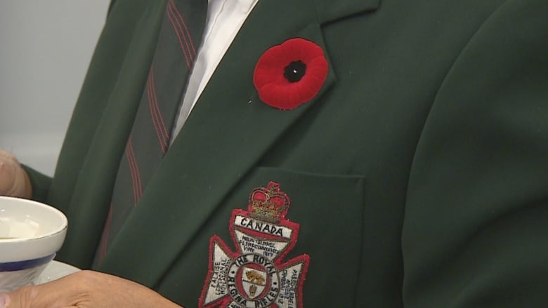 A closeup shows a poppy on the lapel of a man's army-green jacket.