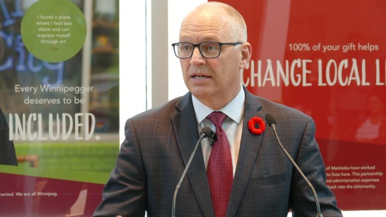 A man is shown speaking at a podium. A poppy pin is seen attached to his suit.