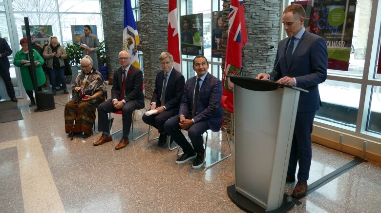 Four people are shown sitting next to a podium, which a man is standing and speaking at.