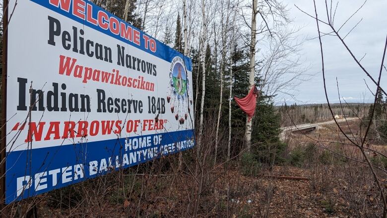 A Pelican Narrows welcome sign overlooking a bridge, with a red dress hung on a nearby tree.