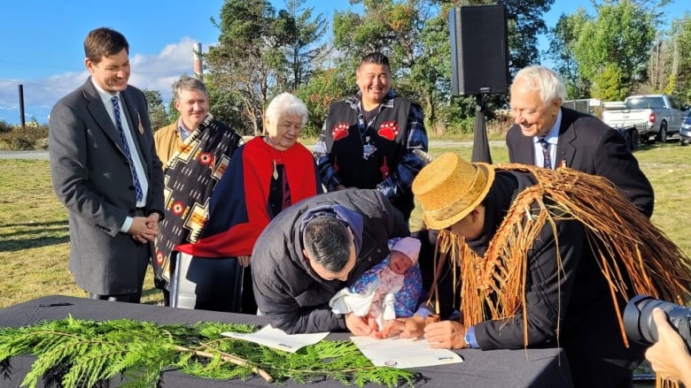 A group of smiling Indigenous adults watch as a father holds his infant's inked foot to a document so she can sign it. 