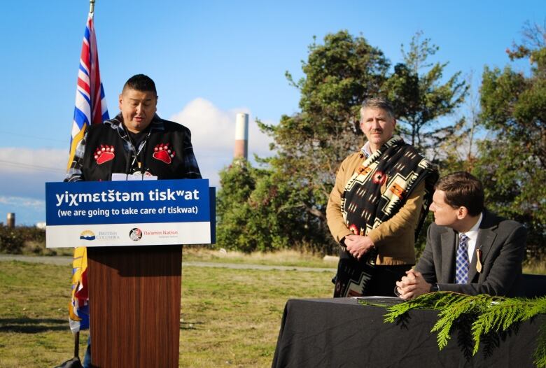 A First Nations man stands at a podium speaking, as two white men look on. Behind them rises the smoke stack of a former mill.