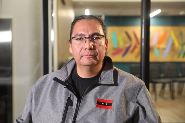 A man in a black shirt and a grey jacket looks directly into the camera. 