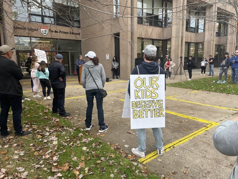 A man at a protest looks toward a speaker and wears a sandwich board with the message 'Our kids deserve better' written across the back. 