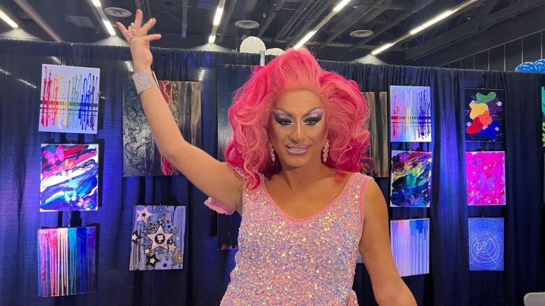 A drag queen in a pink wig and pink sequin dress poses in front of paintings.