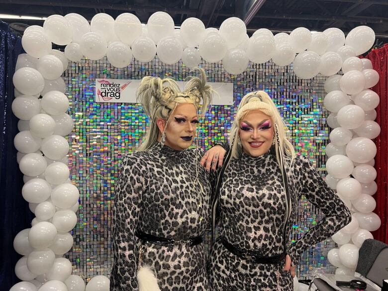 Two drag queens in blonde wigs and leopard print jumpsuits at a convention booth