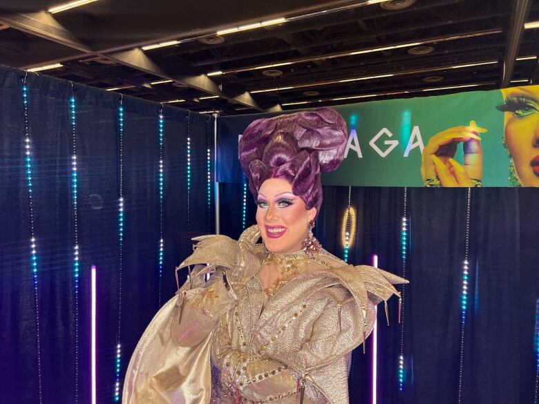 a drag queen with a purple wig and golden dress smiles at the camera