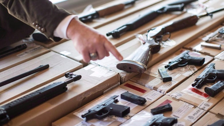 A police officer points at a number of firearms laid out on a table.