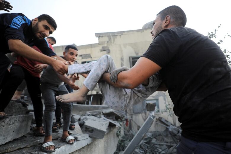 A man standing in a pile of rubble hands a dust-covered child to a group of men waiting nearby. 