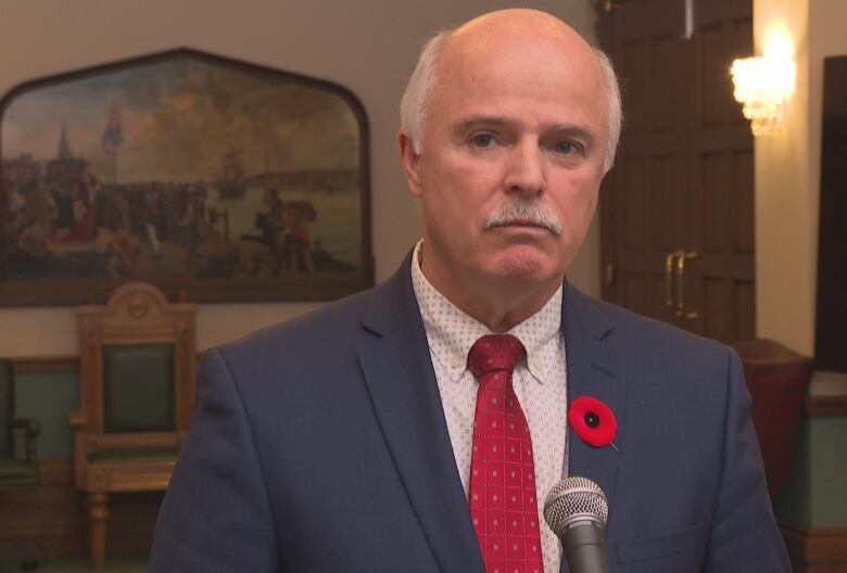 A man in a blue suit and a red tie stands in front of a microphone.