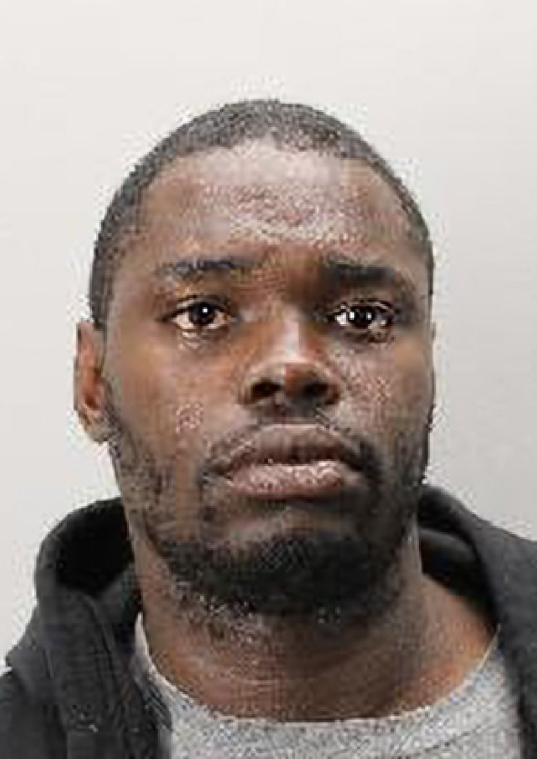 A headshot of a black man with brown eyes and short black hair. He is wearing a grey t shirt and dark grey hoodie.