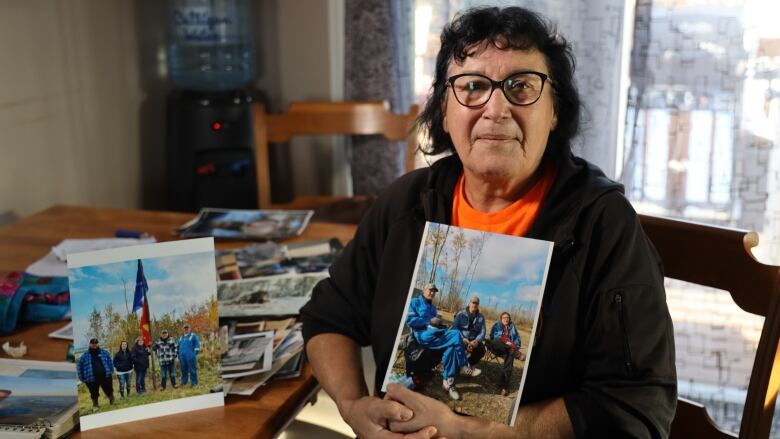 A woman with dark brown hair and glasses, wearing a black hoodie and orange t-shirt, sits at her kitchen table holding a family photograph.