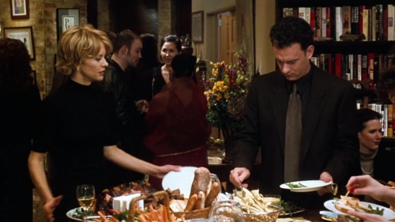 Meg Ryan (left) and Tom Hanks (right) in a still from the movie Youve Got Mail. They're standing at a table of food at a dinner party, putting food on their plates. 